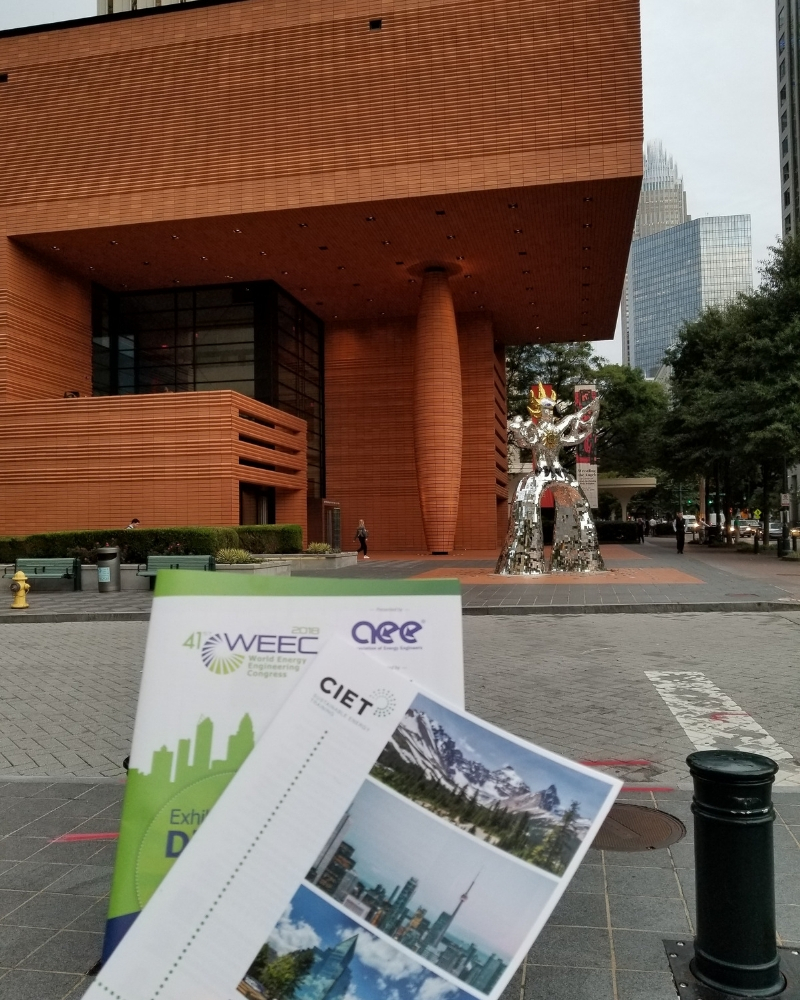 CIET Canada brochure and WEEC brochure being held in front of a metal statue and building in Charlotte, North Carolina